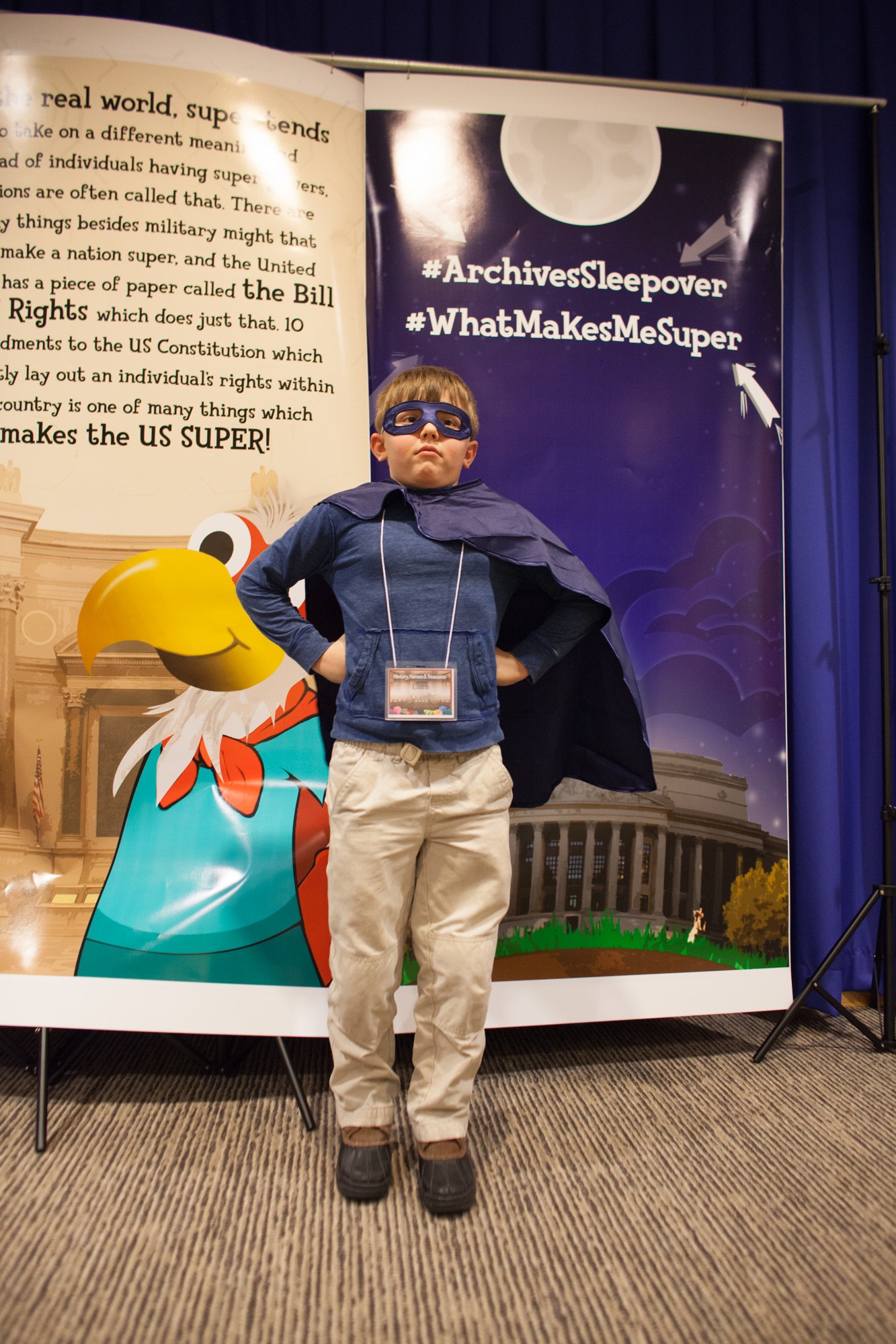 Ein junger Junge in einem Superheldenkostüm steht vor einem Banner mit Text und einem Vogelbild, mit einem Vorhang im Hintergrund, trägt Schuhe.