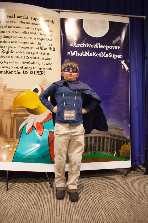 Ein junger Junge in einem Superheldenkostüm steht vor einem Banner mit Text und einem Vogelbild, mit einem Vorhang im Hintergrund, trägt Schuhe.