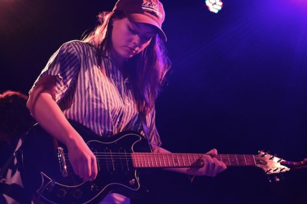 Eine Frau mit Mütze spielt Gitarre auf der Bühne eines Musikfestivals, während zahlreiche Lichter den Hintergrund erhellen.