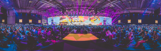 Eine große Menschenmenge, die auf Stühlen sitzt und einer hell erleuchteten Bühne gegenübersteht, umgeben von Bildschirmen, wahrscheinlich auf einem Online-Marketing-Rockstars-Event.