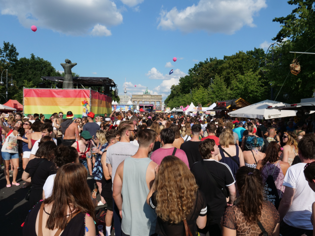 Eine große Menschenmenge, die eine Straße mit Zelten, Bäumen, Pfählen, Lichtern und einer Statue gesäumt entlanggeht, mit Gebäuden und einem bewölkten Himmel mit Ballons im Hintergrund beim Berliner Christopher Street Day.