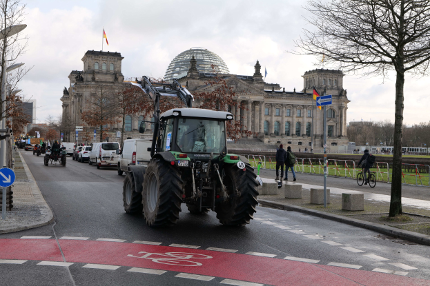 Ein grüner Traktor fährt am Reichstaggebäude in Berlin, Deutschland, vorbei, während Fußgänger und Radfahrer auf dem Bürgersteig unterwegs sind, Bäume die Straße säumen und Fahnen auf dem Gebäude wehen.