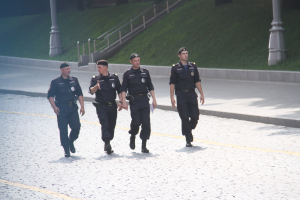 Drei Polizeibeamte in Uniform und Mützen gehen eine Straße entlang mit Pfählen, Geländern, Treppen und Gras im Hintergrund.