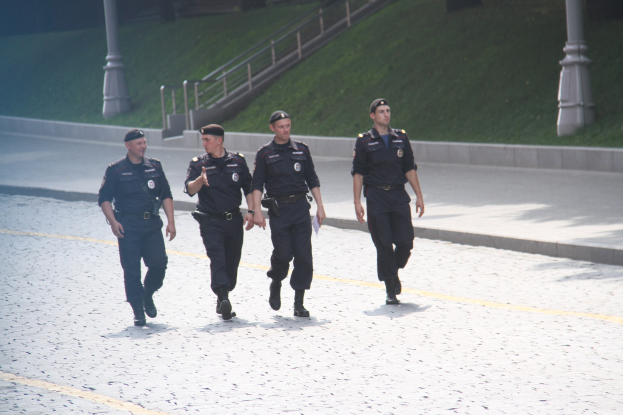 Drei Polizeibeamte in Uniform und Mützen gehen eine Straße entlang mit Pfählen, Geländern, Treppen und Gras im Hintergrund.