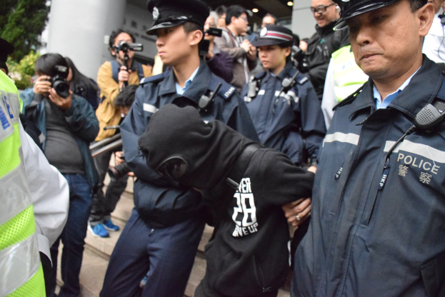 Eine Gruppe von Polizeibeamten steht vor einer Menschenmenge, einige tragen Mützen und halten Kameras, im Hintergrund ein Gebäude mit Säulen und beleuchteter Decke und Bäume links.