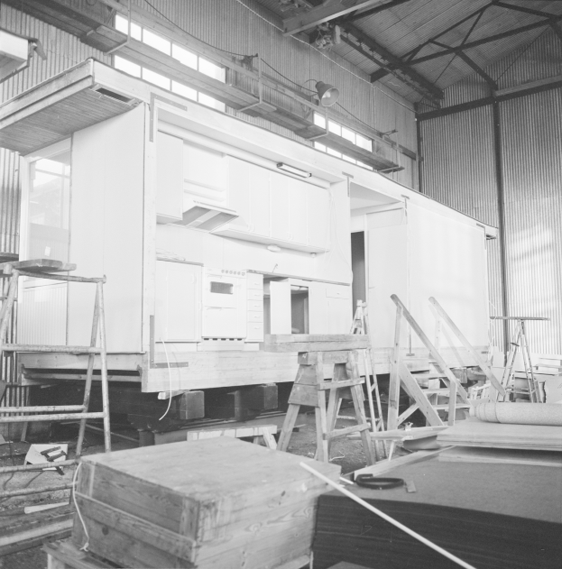 Ein Schwarz-Weiß-Foto eines im Bau befindlichen Gebäudes mit Leitern, Holzboxen und anderen Gegenständen, die auf dem Boden verstreut sind, mit einer Wand mit Fenstern und Lichtern im Hintergrund.