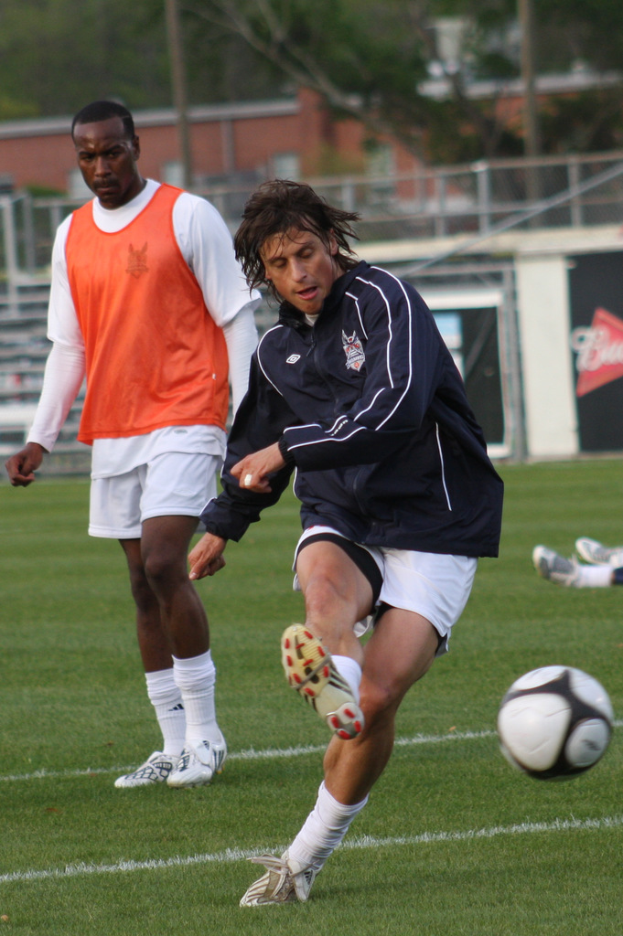 Ein Fußballspieler in einer schwarzen Jacke und weißen Shorts kickt einen Ball, mit einem anderen Spieler in einer orangefarbenen Jacke dahinter und einem eingezäunten Bereich mit Bäumen im Hintergrund.