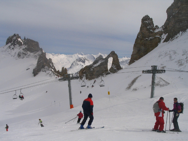 Leute in Pullovern fahren Ski auf Eis, mit Seilbahn, Bergen und bewölktem Himmel im Hintergrund.