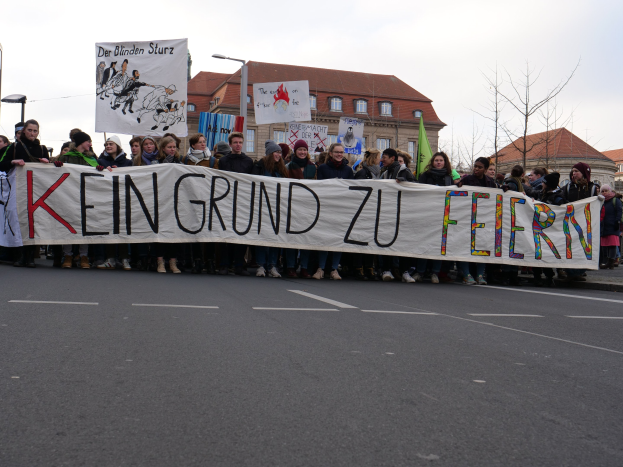 Demonstranten mit einem Transparent "Kein Grund zu Feiern" gegen deutsche Sparpolitik, mit Straßeninfrastruktur und Gebäuden im Hintergrund bei klarem Himmel.