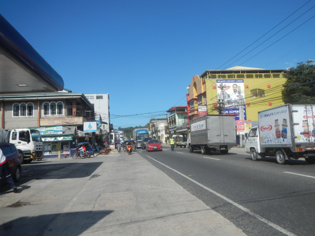 Eine belebte Stadtstraße mit mehreren Autos, Lastwagen, Fußgängern auf dem Gehweg, Gebäuden, Schildern, Strommasten mit Drähten, Bäumen und einem bewölkten Himmel.