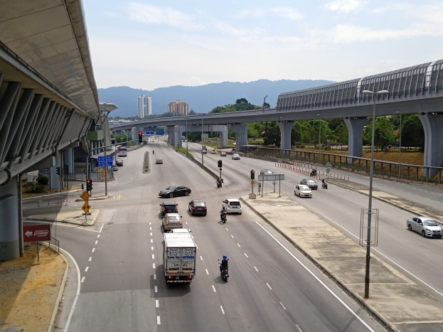 Eine stark befahrene Autobahn mit Schwerlastverkehr neben einer Brücke, mit Straßenlaternen, Verkehrsampeln, Verkehrszeichen, Bäumen, Gebäuden, Hügeln und einem bewölkten Himmel im Hintergrund.