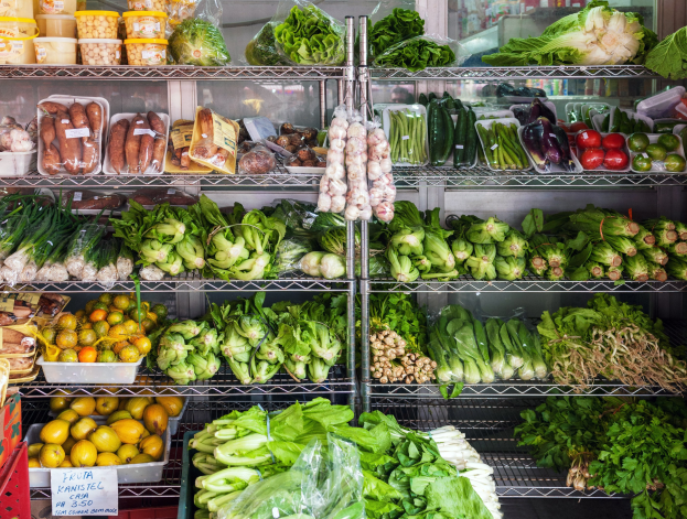 Ein Gang in einem Supermarkt mit frischem Obst und Gemüse in Plastikverpackungen, Kisten und einer Tafel, sowie einem Glasfenster im Hintergrund.