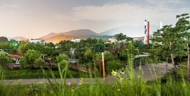 Ein Park mit grünem Gras, bunten Blumen, hohen Bäumen, einer Brücke, einem Springbrunnen, Laternenpfählen und Gebäuden im Hintergrund, vor Bergen und einem klaren blauen Himmel.