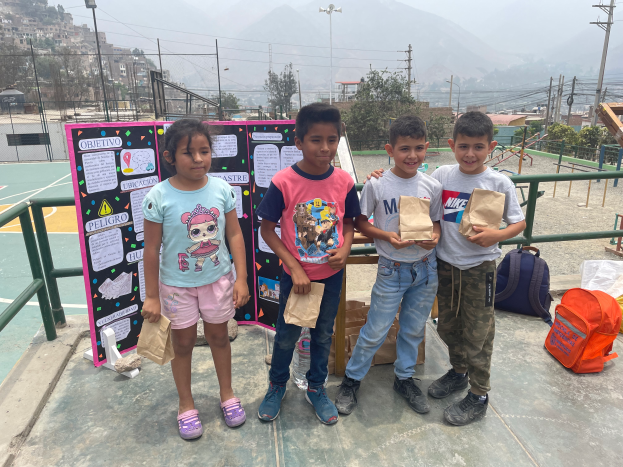 Eine Gruppe von Kindern steht auf einem Basketballfeld, jedes hält einen Papiertüte in der Hand, mit Tüten und Gegenständen rechts daneben und einer Tafel mit Text links daneben, mit Zaun, Pfosten, Straßenlaternen, Bäumen, Gebäuden, Hügeln und Himmel im Hintergrund.