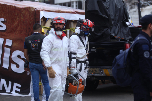 Eine Gruppe von Menschen in Schutzausrüstung, einschließlich Helmen, die eine Straße entlanggehen, mit einem Lastwagen und Gebäuden im Hintergrund, eine Person hölt ein Objekt, und eine Fahne mit Text auf der linken Seite.