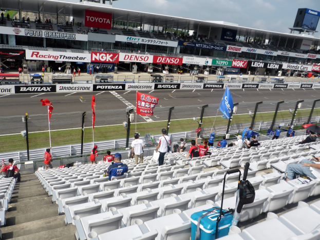 Eine Gruppe von Menschen auf Tribünen bei einer Rennstrecke mit Fahnen, Texttafeln, Geländern und Stufen, mit einem bewölkten Himmel über einem Stadion im Hintergrund.