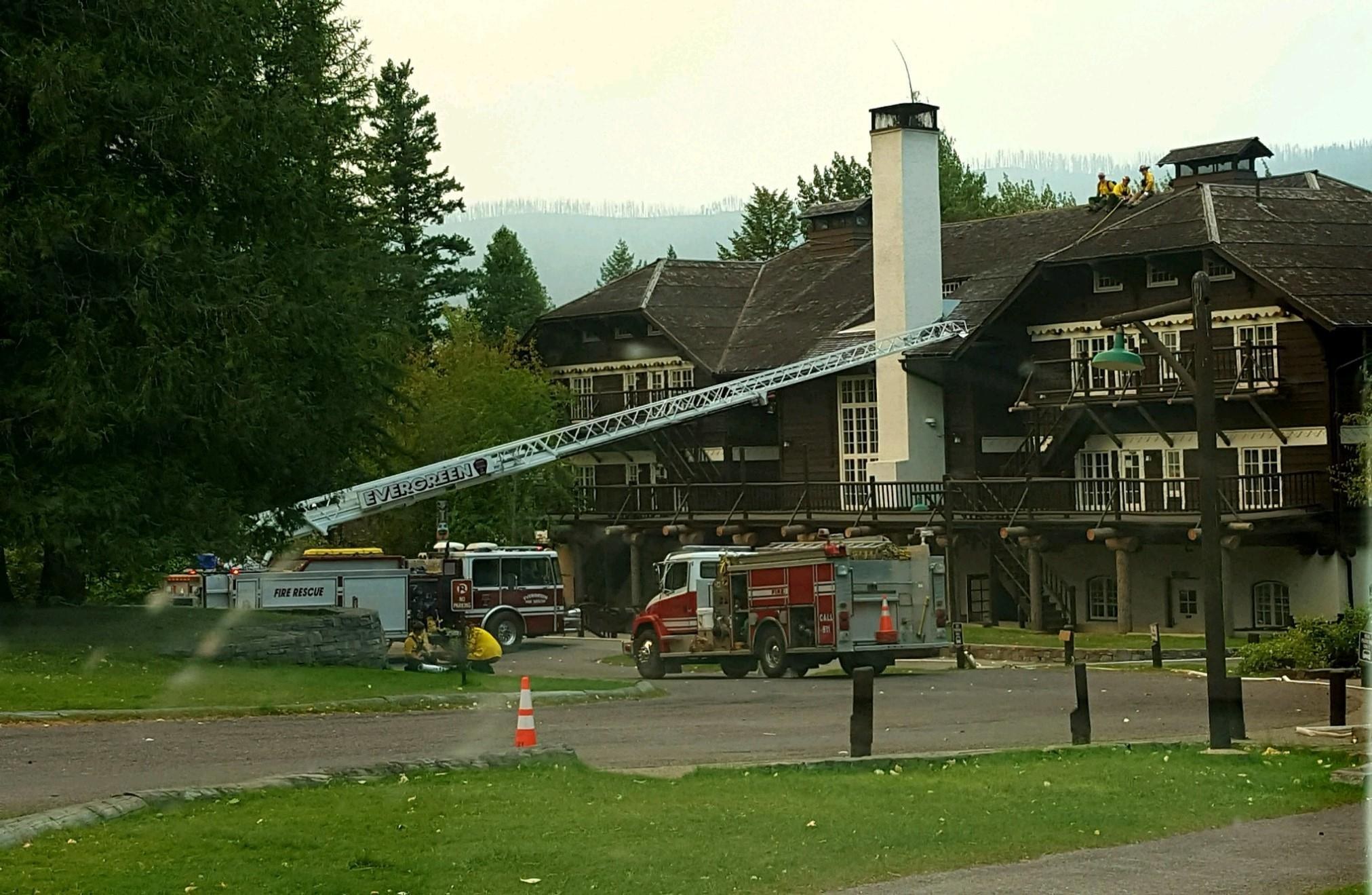 Feuerwehrfahrzeug vor einem großen Gebäude mit Fenstern, Geländern, Säulen und einem Schornstein geparkt, umgeben von Absperrbaken, Gras, Bäumen und Laternen, mit ein paar Menschen in der Nähe, Hügel im Hintergrund und einem klaren blauen Himmel.