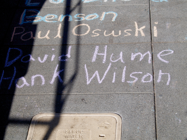Ein Gehweg mit Kreide-Schrift, die wie ein Denkmal aussieht, ein Gullydeckel am Boden und ein Baumschatten auf der linken Seite.