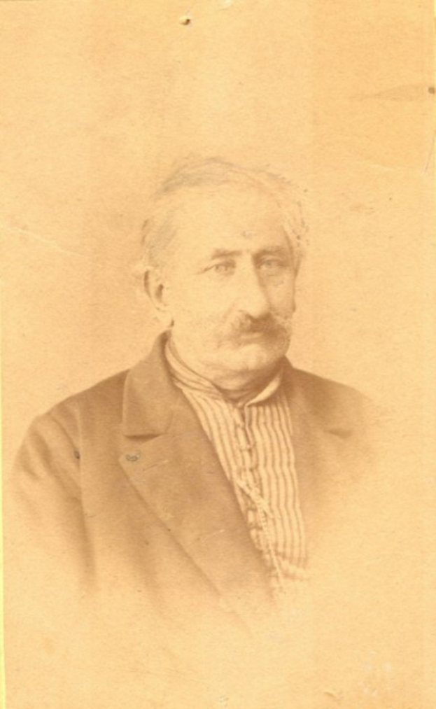 Ein altes Schwarz-Weiß-Porträt eines älteren Mannes mit Schnurrbart, der einen einfachen Mantel trägt und ein starkes Kinn, eine prominente Nase, dunkle Augen und einen nachdenklichen Ausdruck hat.