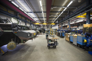 Ein großes Lagerinnere mit Maschinen, Fahrzeugen auf der linken Seite, Transportwagen mit Gegenständen, Holzboxen und anderen Gegenständen auf der rechten Seite und Deckenbeleuchtung.