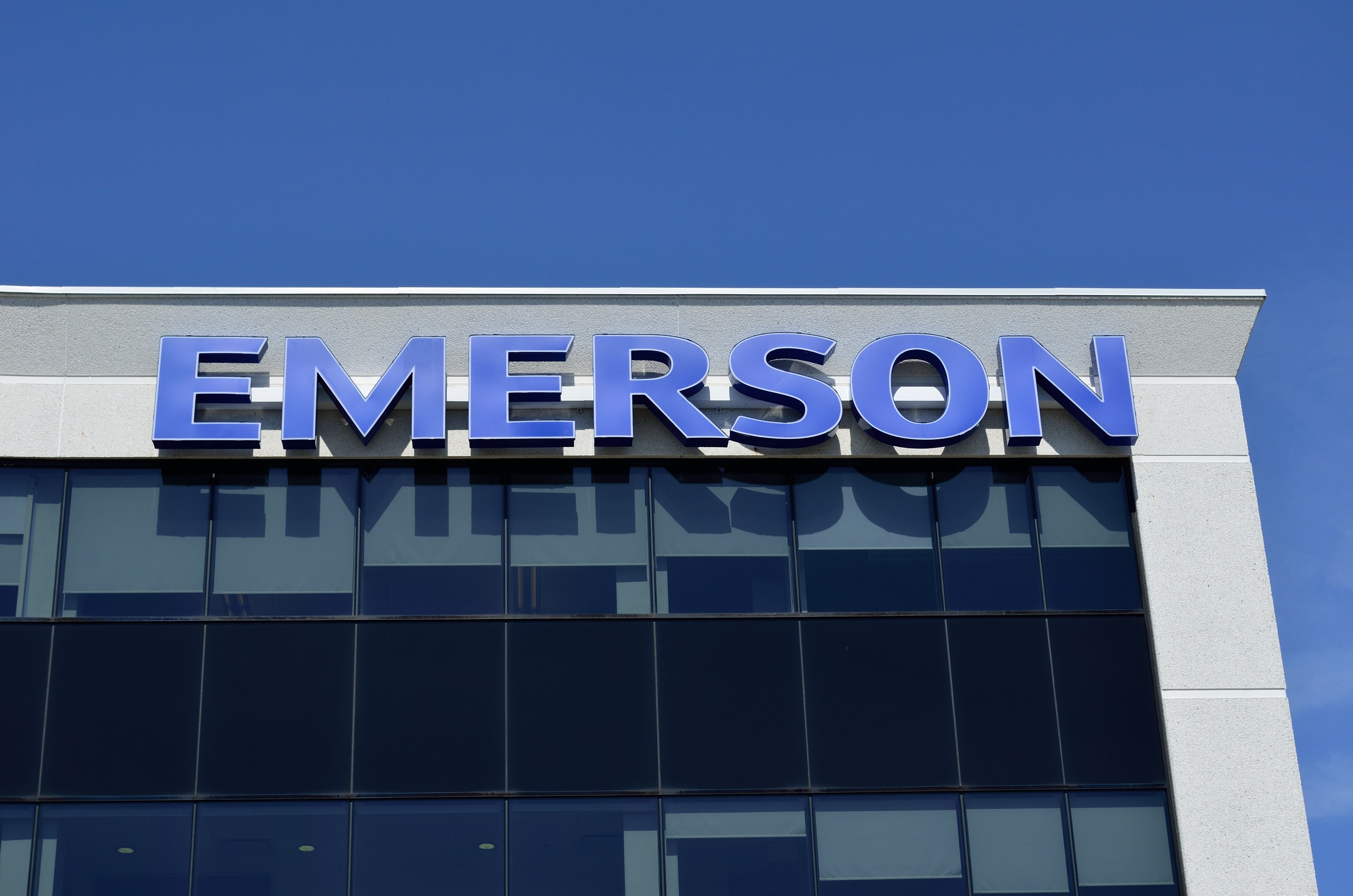 Außenansicht eines Emerson-Gebäudes mit dem Firmenlogo und dem Text "Emerson investiert 1,5 Milliarden USD in den USA" vor einem klaren blauen Himmel.