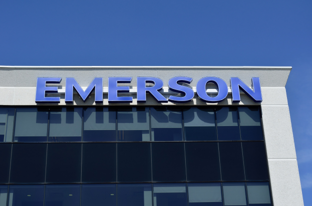 Außenansicht eines Emerson-Gebäudes mit dem Firmenlogo und dem Text "Emerson investiert 1,5 Milliarden USD in den USA" vor einem klaren blauen Himmel.