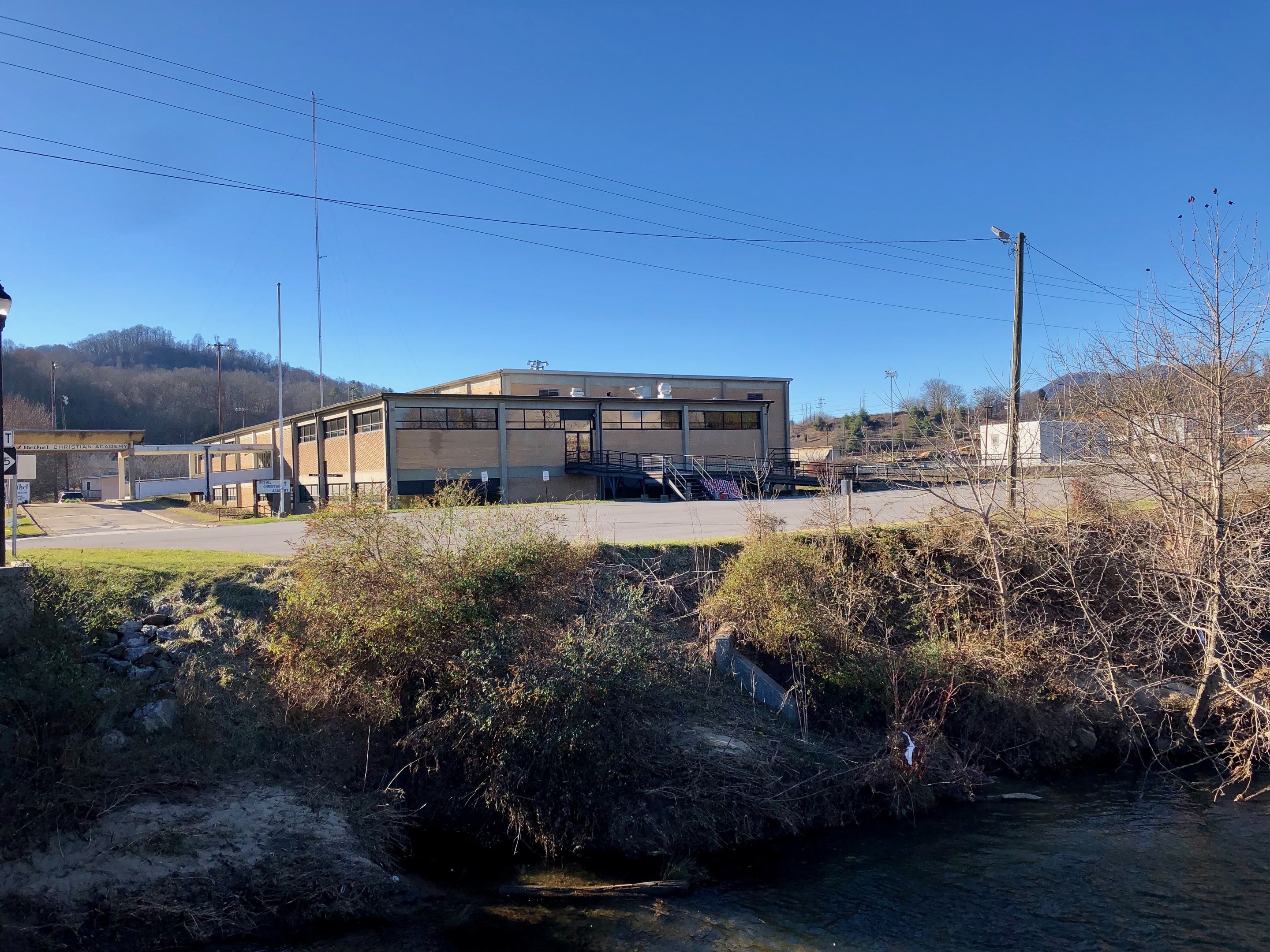 Grünes Feld mit einem Fluss, B├Ąumen, Pflanzen, Gras, Steinen, Stra├čenlaternen, Strommasten, Stromkabeln, H├Ąngen, einem klaren blauen Himmel und einem Geb├Ąude an der Stelle einer geplanten Wasseraufbereitungsanlage.