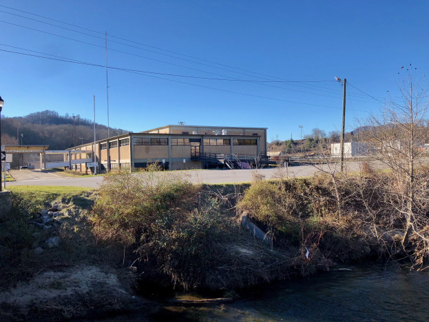 Grünes Feld mit einem Fluss, B├Ąumen, Pflanzen, Gras, Steinen, Stra├čenlaternen, Strommasten, Stromkabeln, H├Ąngen, einem klaren blauen Himmel und einem Geb├Ąude an der Stelle einer geplanten Wasseraufbereitungsanlage.