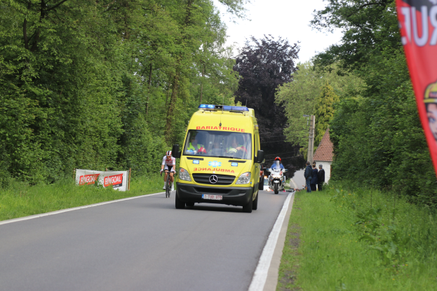 Ein Rettungswagen f√§hrt auf einer Stra√Ÿe mit Fahrradfahrern daneben, umgeben von Gras, B√§umen, H√§usern, Masten und einem klaren blauen Himmel.