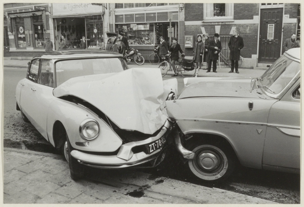 Schwarz-weiß-Foto von zwei beschädigten Autos am Straßenrand mit Menschen und Fahrrädern in der Nähe und Gebäuden im Hintergrund.
