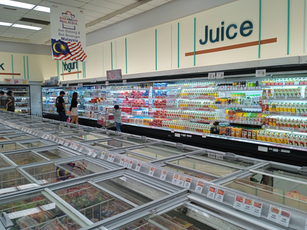Ein hell erleuchteter Supermarktgang mit Flaschengetränken, frischem Obst und anderen Lebensmitteln, sowie einigen Kunden, die durch die Gänge schlendern und einer beleuchteten Anzeigetafel an der Decke.