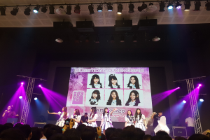 Eine Gruppe von Menschen performt auf einer hell erleuchteten Bühne mit Trägern, während ein Sänger ein Mikrofon hält und ein anderer ein Instrument spielt, während die Menge darunter das Konzert aufnimmt; ein großer Bildschirm im Hintergrund zeigt Bilder der K-Pop-Gruppe BK48.