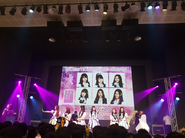 Eine Gruppe von Menschen performt auf einer hell erleuchteten Bühne mit Trägern, während ein Sänger ein Mikrofon hält und ein anderer ein Instrument spielt, während die Menge darunter das Konzert aufnimmt; ein großer Bildschirm im Hintergrund zeigt Bilder der K-Pop-Gruppe BK48.