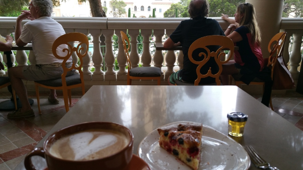 Ein älterer Mann sitzt an einem Tisch mit einer Kaffeetasse, einem Teller mit Essen und einer Gabel.
