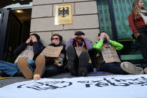 Eine Gruppe von Menschen sitzt vor einem Gebäude auf dem Boden und hält Schilder, mit einer Fahne und einem Apfel auf der Straße, und eine Frau steht rechts daneben und sieht besorgt aus.