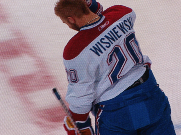 Ein Hockey-Spieler in einem "Wisniewski"-Trikot steht auf dem Eis und hält einen Hockey-Schläger, mit einem konzentrierten und entschlossenen Gesichtsausdruck.
