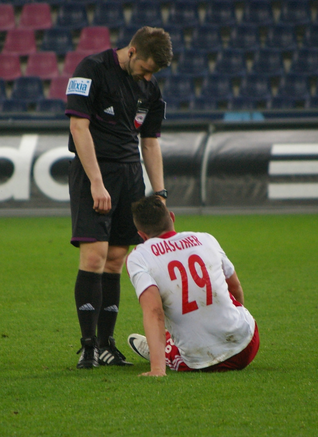 Ein Fussballspieler sitzt neben einem Schiedsrichter auf dem Boden, beide in Sportkleidung, mit Tribünen und Stühlen im Hintergrund.