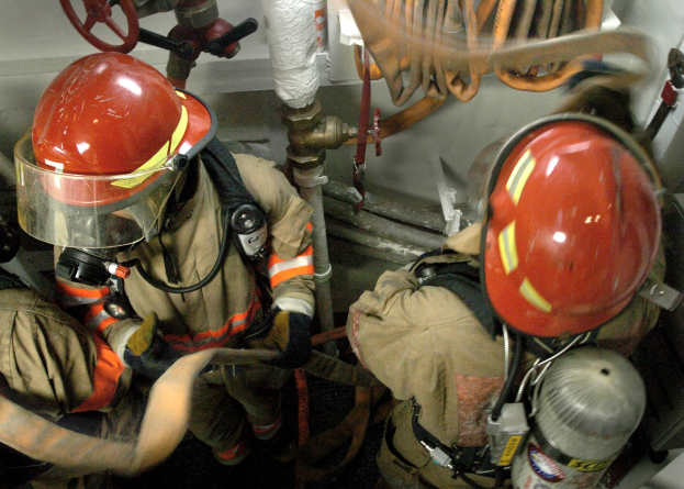 Feuerwehrleute in Schutzausrüstung bei der Arbeit an einem Feuerhydranten mit Rohren und Ventilen im Hintergrund.