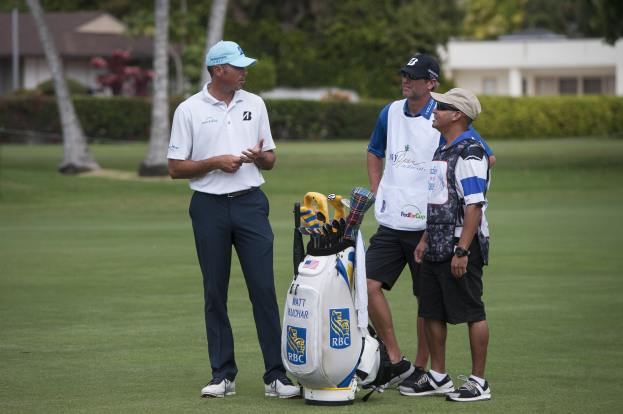Mannergruppe auf einem grünen Feld mit einem Golfbag im Vordergrund, Bäumen, Pflanzen und Gebäuden im Hintergrund.