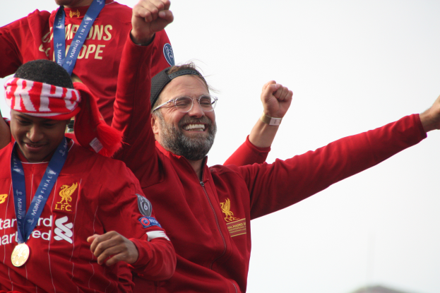 Liverpool-Trainer Jürgen Klopp im roten Jackett feiert mit seinen Spielern nach dem Gewinn der UEFA Champions League, während zwei medaillentragende Personen in der Nähe sind und der Himmel im Hintergrund zu sehen ist.