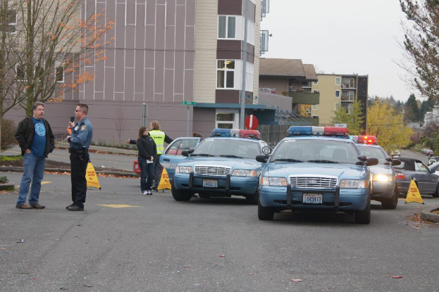Autos auf einer Straße mit vier Menschen in der Nähe, Gebäude mit Fenstern im Hintergrund, Bäume und ein Warndreieck.