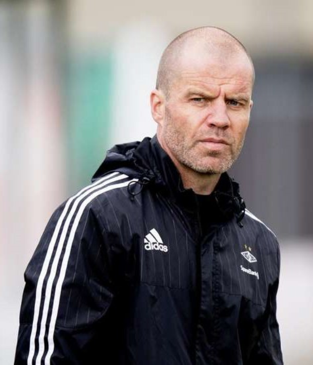 Ein kahlköpfiger Trainer in einer blauen Jacke mit weißen Streifen und einem weißen Kragen steht auf einem Fußballfeld und schaut ernst nach vorne, die Hände vor der Brust verschränkt.