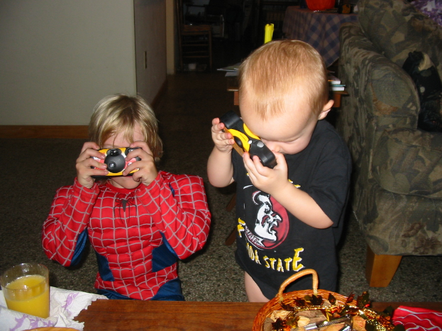 Zwei Kinder, eines im Spider-Man-Kostüm, halten eine Spielkamera, mit einem Holztisch, Glas und Korb im Vordergrund und einem Sofa und einem weiteren Holztisch im Hintergrund.