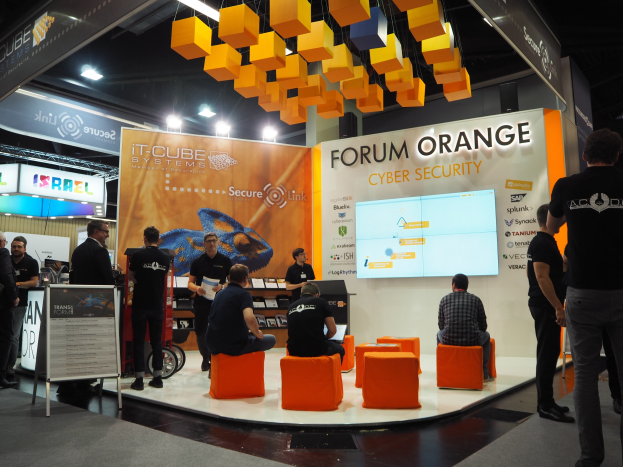 Eine Gruppe von Menschen an einem IT-Cube Systems Forum Orange Cyber Security Stand auf einer Messe, mit sitzenden und stehenden Teilnehmern, einer Tafel mit Text, einem Anzeigebildschirm, Regalen mit Gegenständen, Schildern, Deckenleuchten und Lampen.