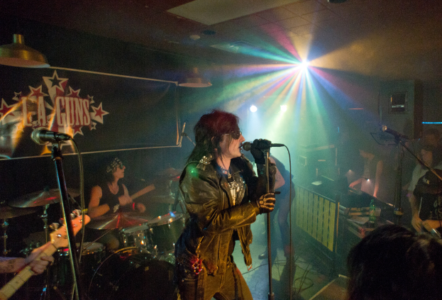 Eine Frau in einer schwarzen Jacke singt in ein Mikrofon auf der Bühne, mit einem Mann in Sonnenbrille und Headband, der ein Instrument spielt, und einem Plakat mit 'LA Guns' im Hintergrund.