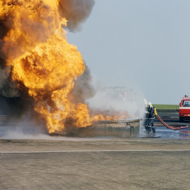Feuerwehrlöschfahrzeug in Flammen auf der Straßenseite mit zwei Helmen tragenden Personen, die Rohre halten, einem Fahrzeug im Hintergrund und dem Himmel.