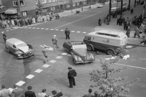 Schwarz-weiß-Szene eines Autounfalls an einer Straßenkreuzung mit einem Lieferwagen, anderen Fahrzeugen und einer Gruppe von Menschen; Gebäude, Bäume, Pfosten und Texttafeln im Hintergrund.