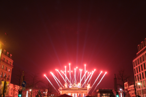 Eine nächtliche Stadtstraße mit Gebäuden, Bäumen, Laternen, Verkehrsampeln, Schildern und Menschen, mit Feuerwerk, das den Himmel erleuchtet.