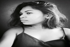 Eine selbstbewusste Frau in einem schwarzen Kleid posiert in einem Schwarz-Weiß-Foto vor einer Wand mit einem glatten Bob und natürlichem Make-up.