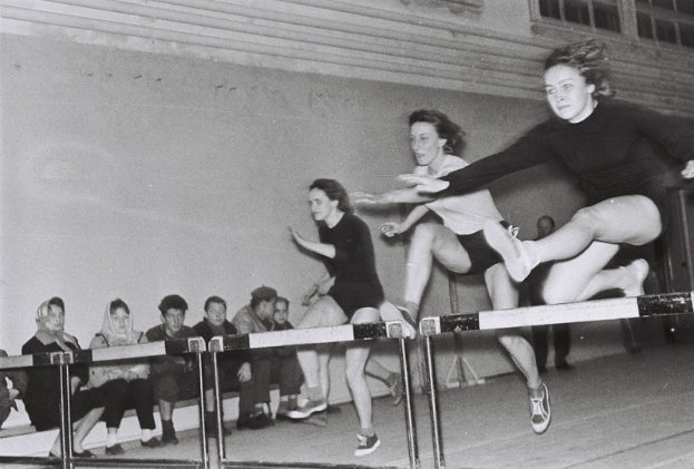 Zwei Frauen in der Luft, über ein Hindernis springend in einer Turnhalle, mit Zuschauern auf der linken Seite und einer Wand mit Fenstern im Hintergrund.